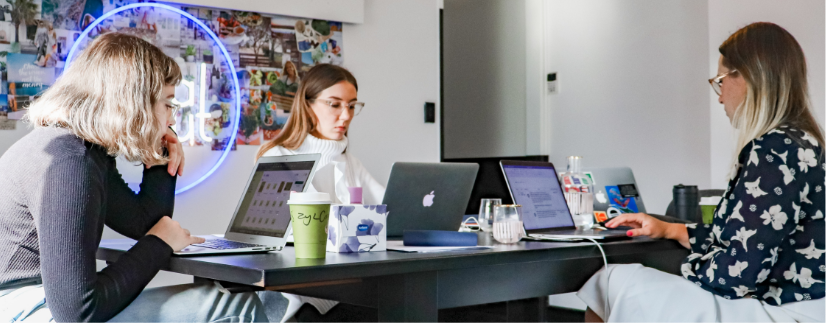 Three people sitting at a long table with their laptops open. 