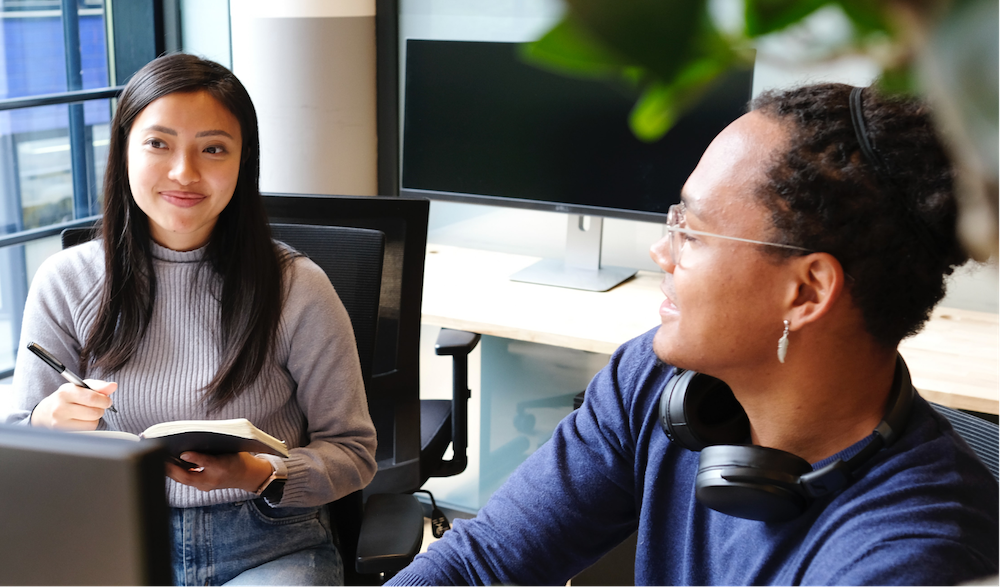 Two people sitting at a disk in front of a monitor having a conversation. 